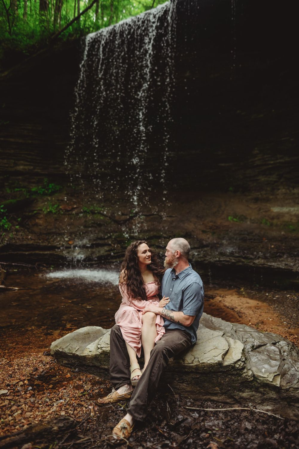 A couple sits together on rocks beneath a serene waterfall in a forest setting.