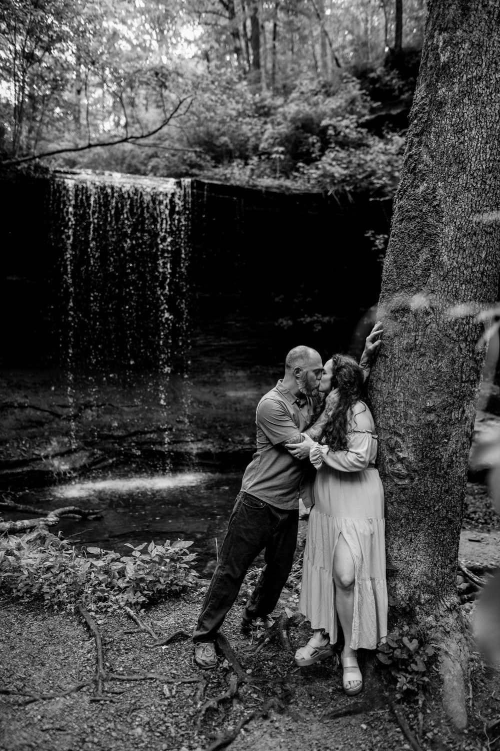 A romantic black and white portrait by a waterfall with a couple embracing against a tree.