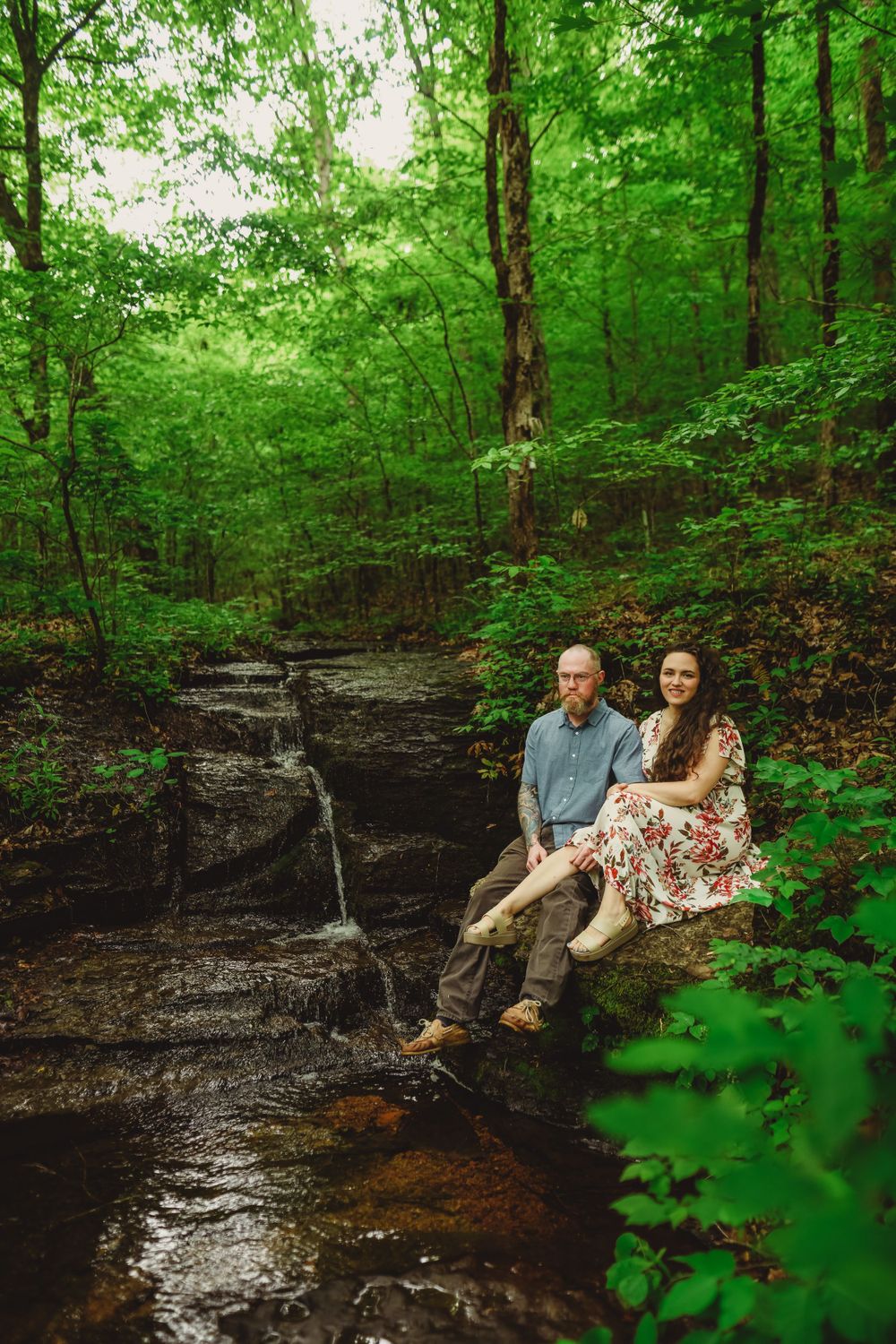 A couple relaxes on dark rocks near a forest stream surrounded by vibrant green foliage.