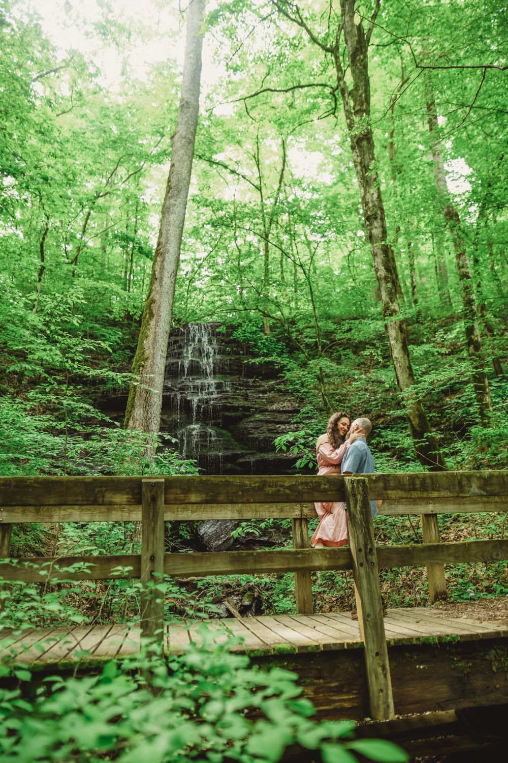 A couple stands on a wooden boardwalk viewing a cascading waterfall in a lush green forest setting.