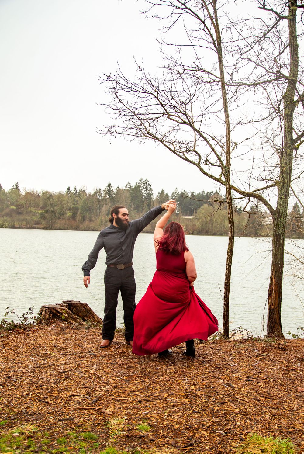 A couple dances near a lake with bare winter trees in the background.