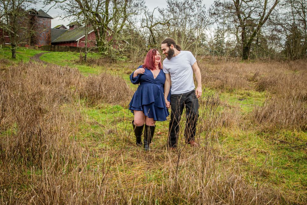 A couple walking together through a grassy field with a barn visible in the background.