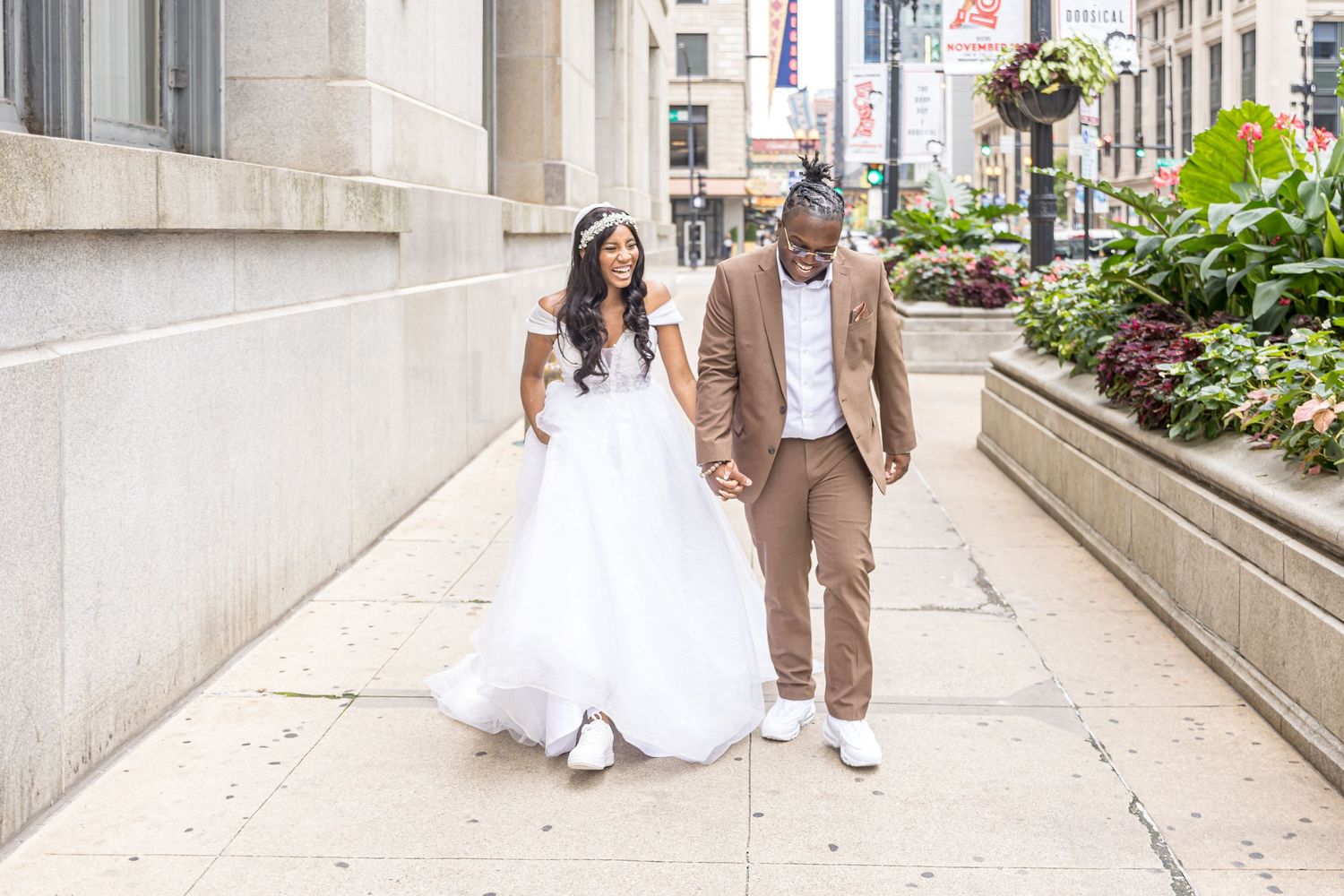 A smiling bride and groom stroll down a city sidewalk, with her in white and him in a tan suit.