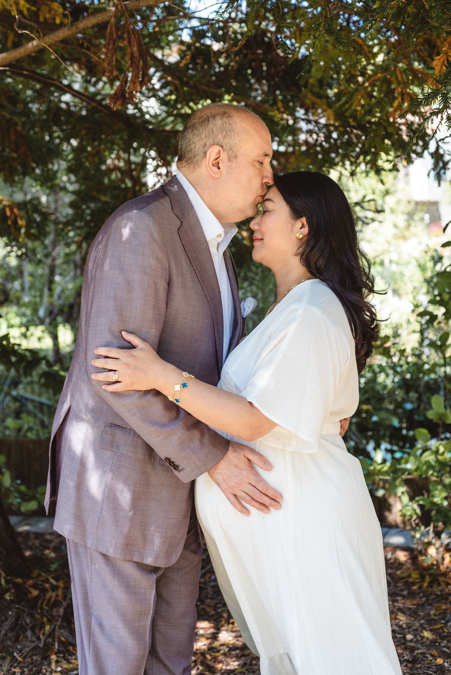 A romantic couple embraces outdoors under trees, with the person in a grey suit and other in white dress.