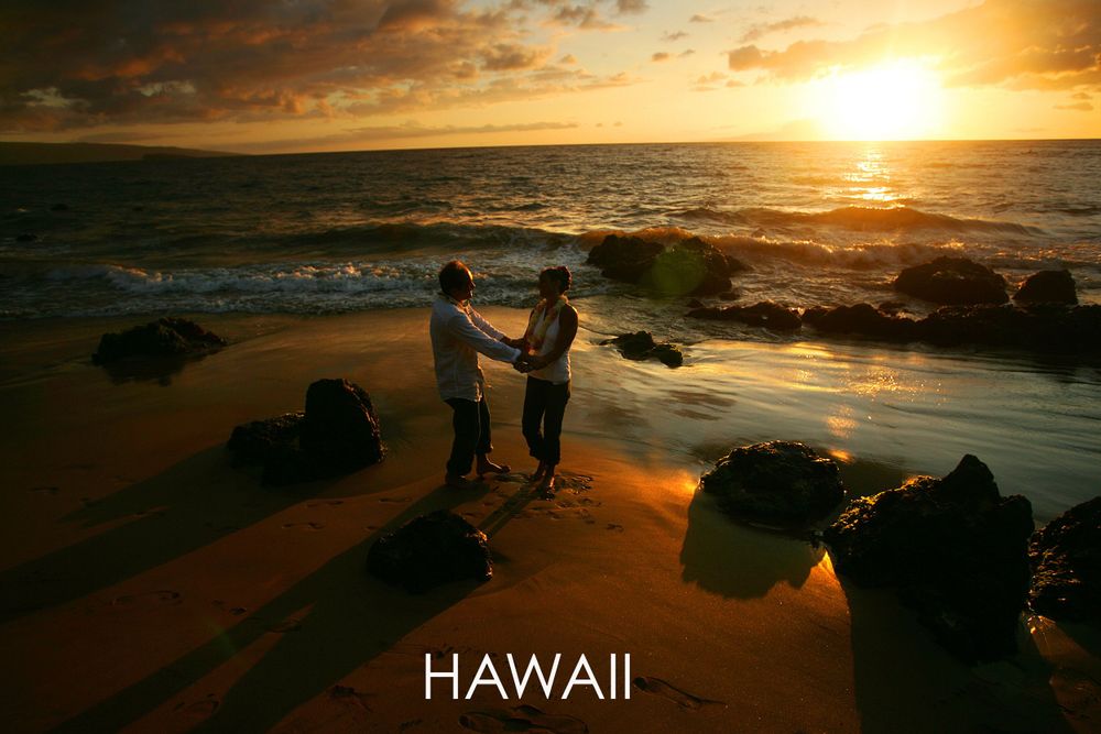 Silhouetted couple holding hands on rocky beach during golden Hawaiian sunset.