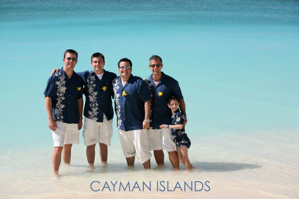 Group wearing Hawaiian shirts poses on white sand beach with turquoise waters in Cayman Islands.
