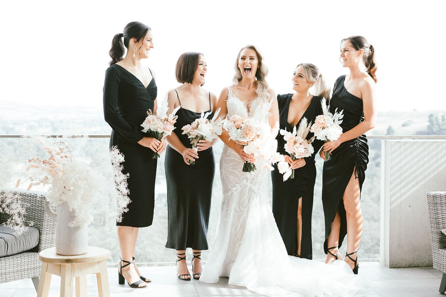 Wedding party in coordinated black dresses and white bouquets laugh together during formal photos.