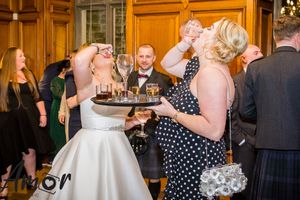 Wedding reception scene showing guests enjoying drinks and celebrations in a wood-paneled room.