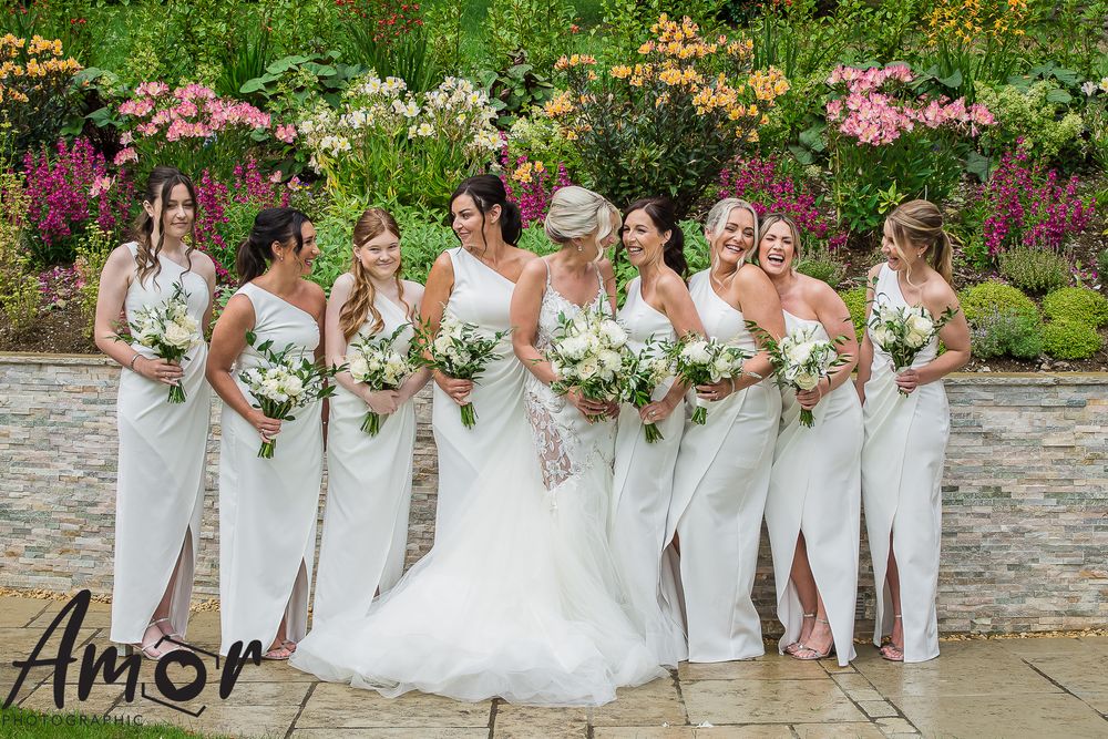 Bridal party in white dresses poses against colorful garden backdrop.
