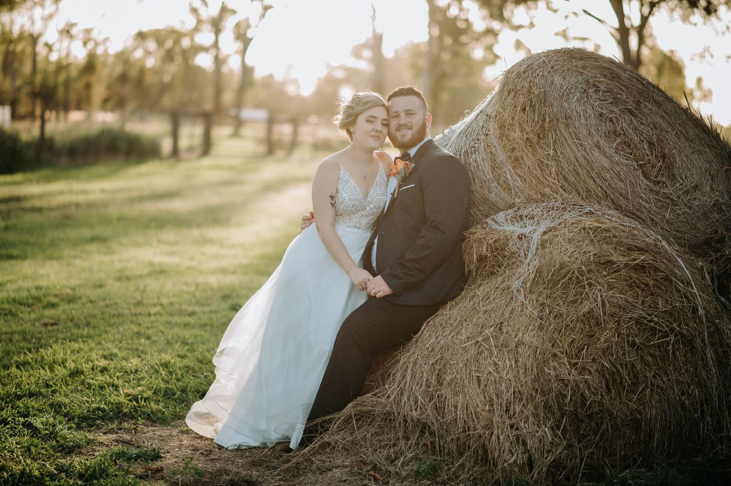 Newlyweds posing together against rustic hay bale in golden afternoon light.