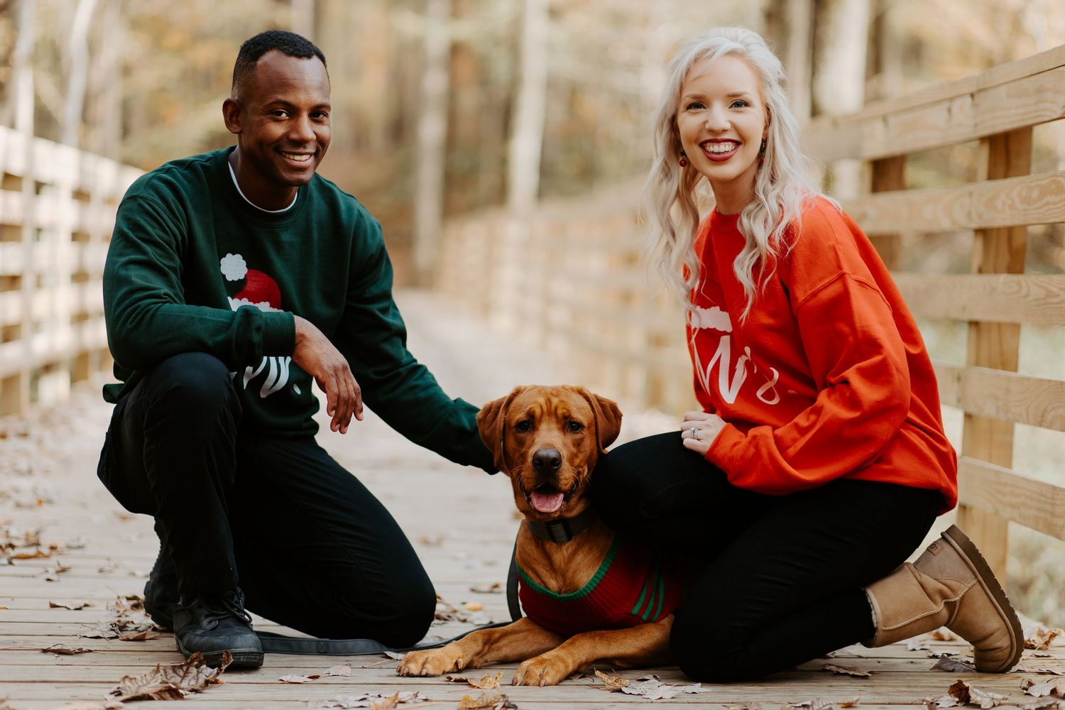 Smiling couple in holiday sweaters kneeling with their brown Vizsla dog on a wooden bridge outdoors.