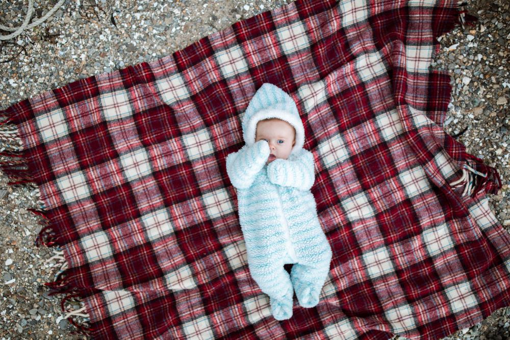 Baby in striped blue snowsuit lying on red plaid blanket in winter setting.