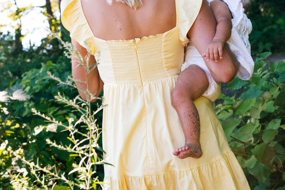 Back view of someone in a yellow sundress holding a baby outdoors.