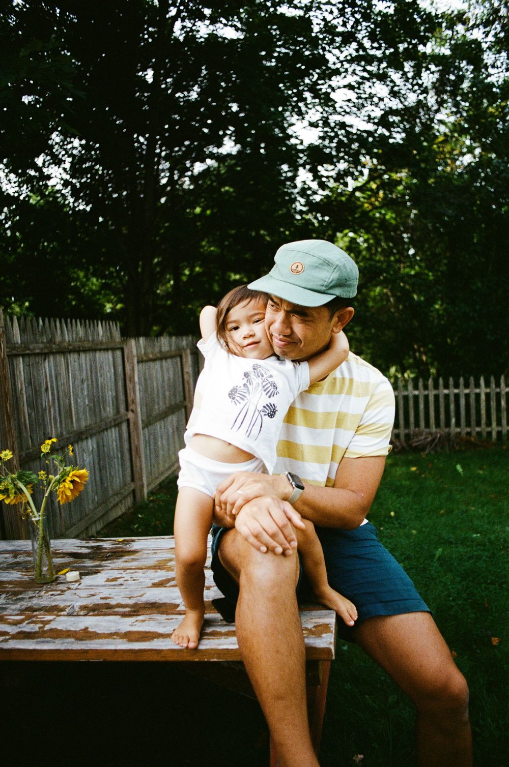 A tender moment shared between parent and child sitting outdoors on a wooden surface in a backyard garden setting.