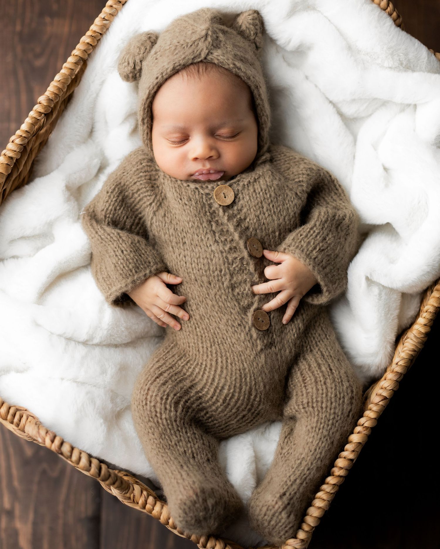 Newborn baby wearing cozy brown knit outfit sleeping in wooden basket with white padding.