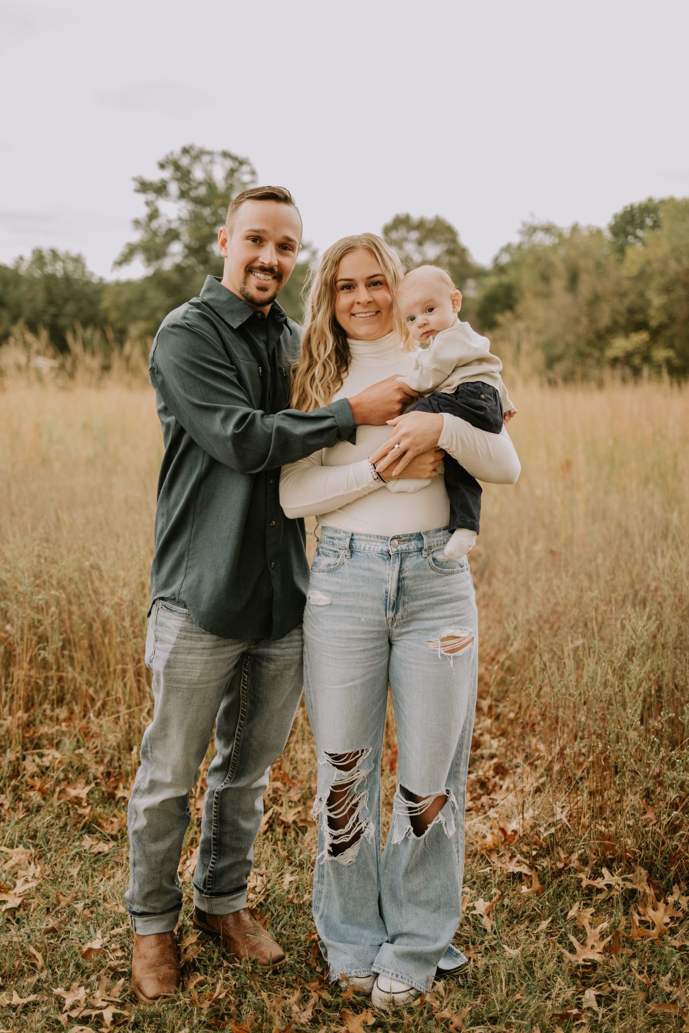 A young family poses together in a sunlit autumn field wearing casual clothes.