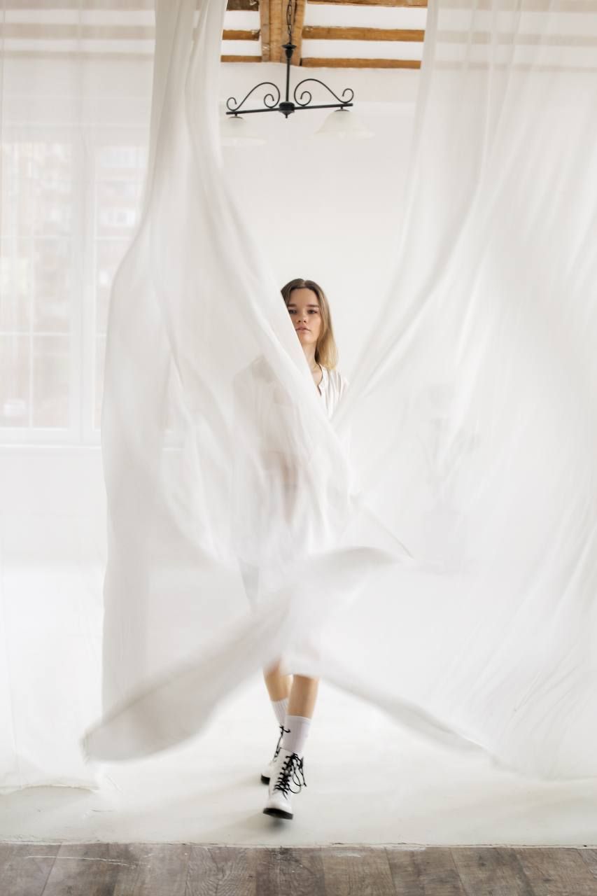Figure draped in flowing white fabric poses against curtains with dramatic movement.