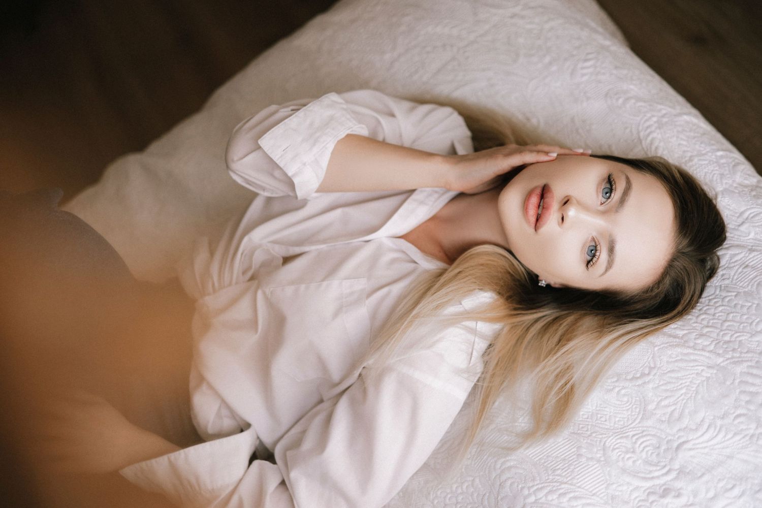 Person lying on white bedding wearing light colored shirt with serene expression in soft lighting.