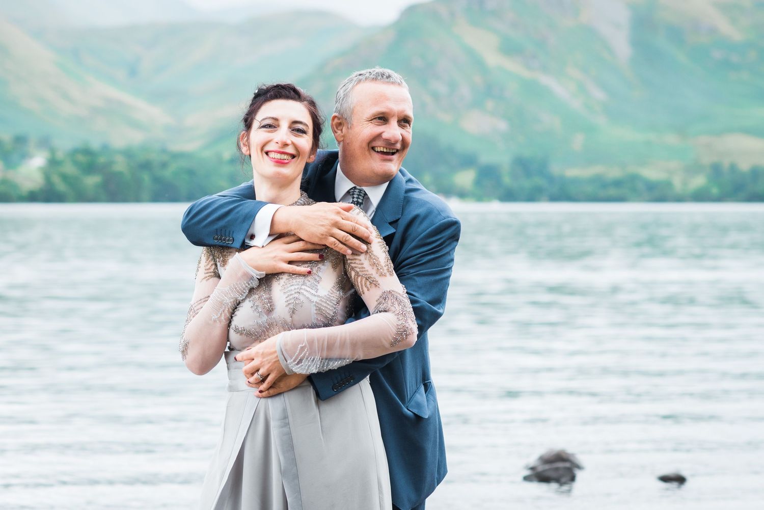 Couple embrace infant of Ullswater Lake with the view of mountains behind.