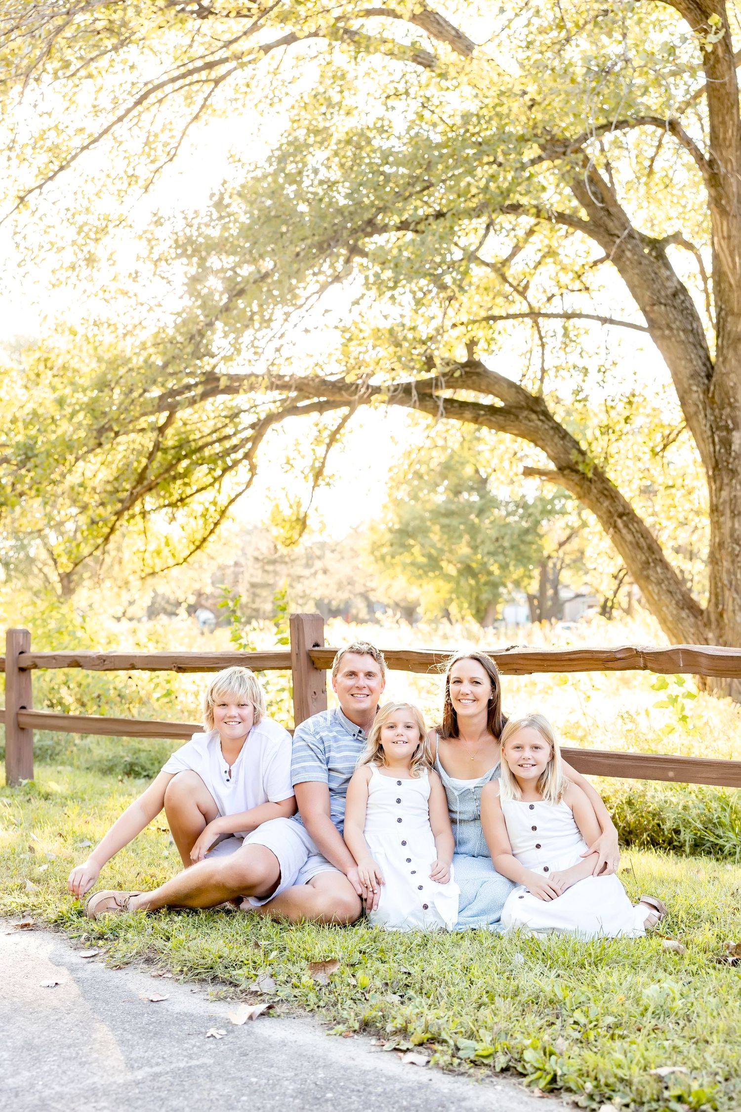 Family relaxes together on grassy hill near wooden fence during golden hour photo session.