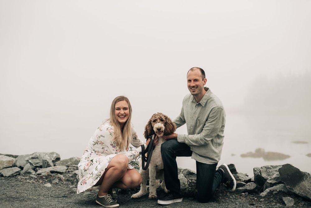 A couple sits with their fluffy dog on rocky terrain surrounded by fog.