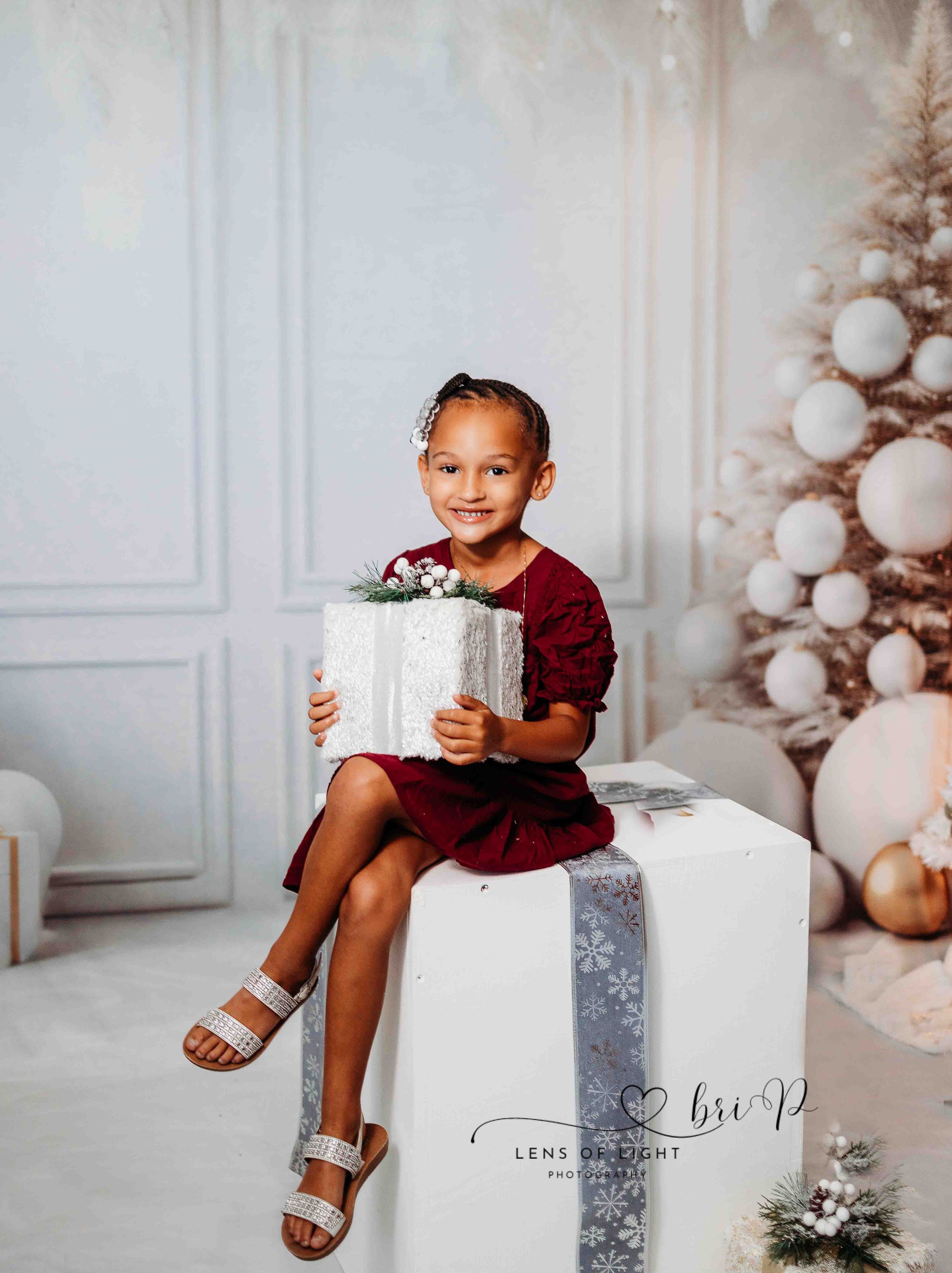 A child in a burgundy dress sits with a silver present against a winter holiday backdrop.