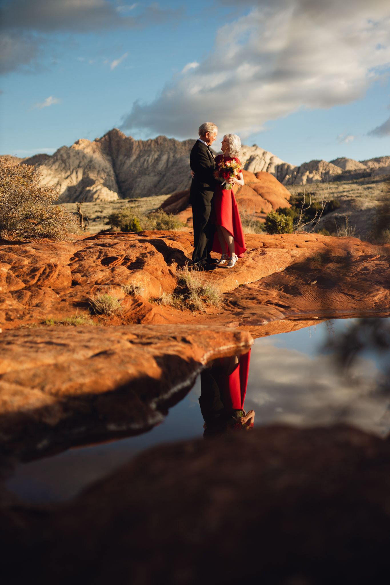 Senior couple embracing on red rocks reflected in a still pool with mountain backdrop at golden hour.