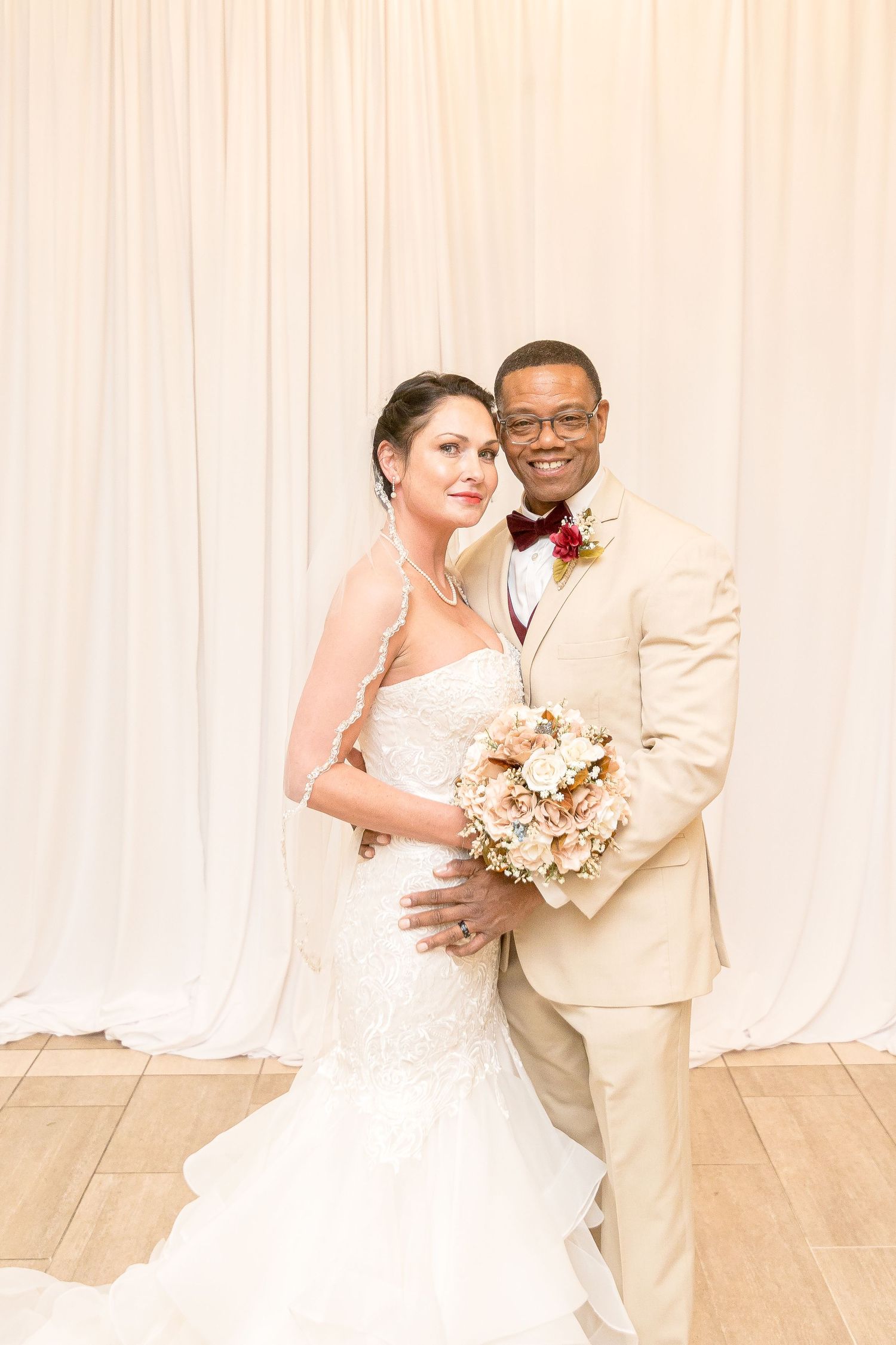 A happy couple poses for their wedding portrait in cream and white attire with a beautiful floral bouquet.