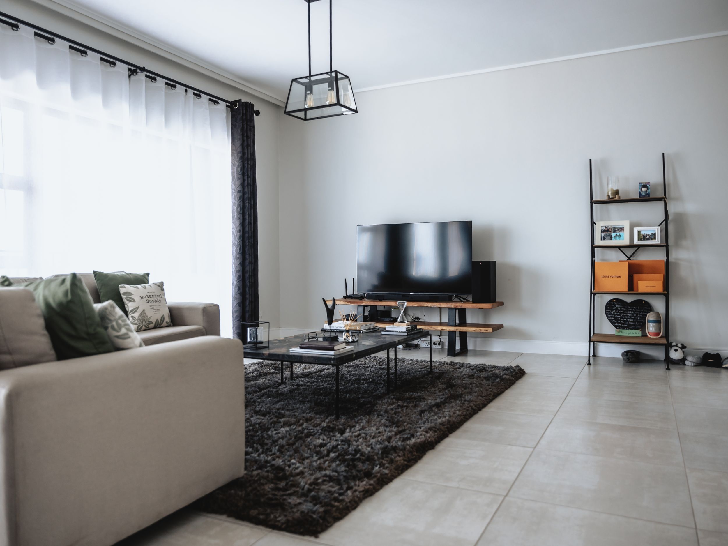 Modern living room with beige sectional sofa, black rug, TV unit and ladder shelf in minimalist style.