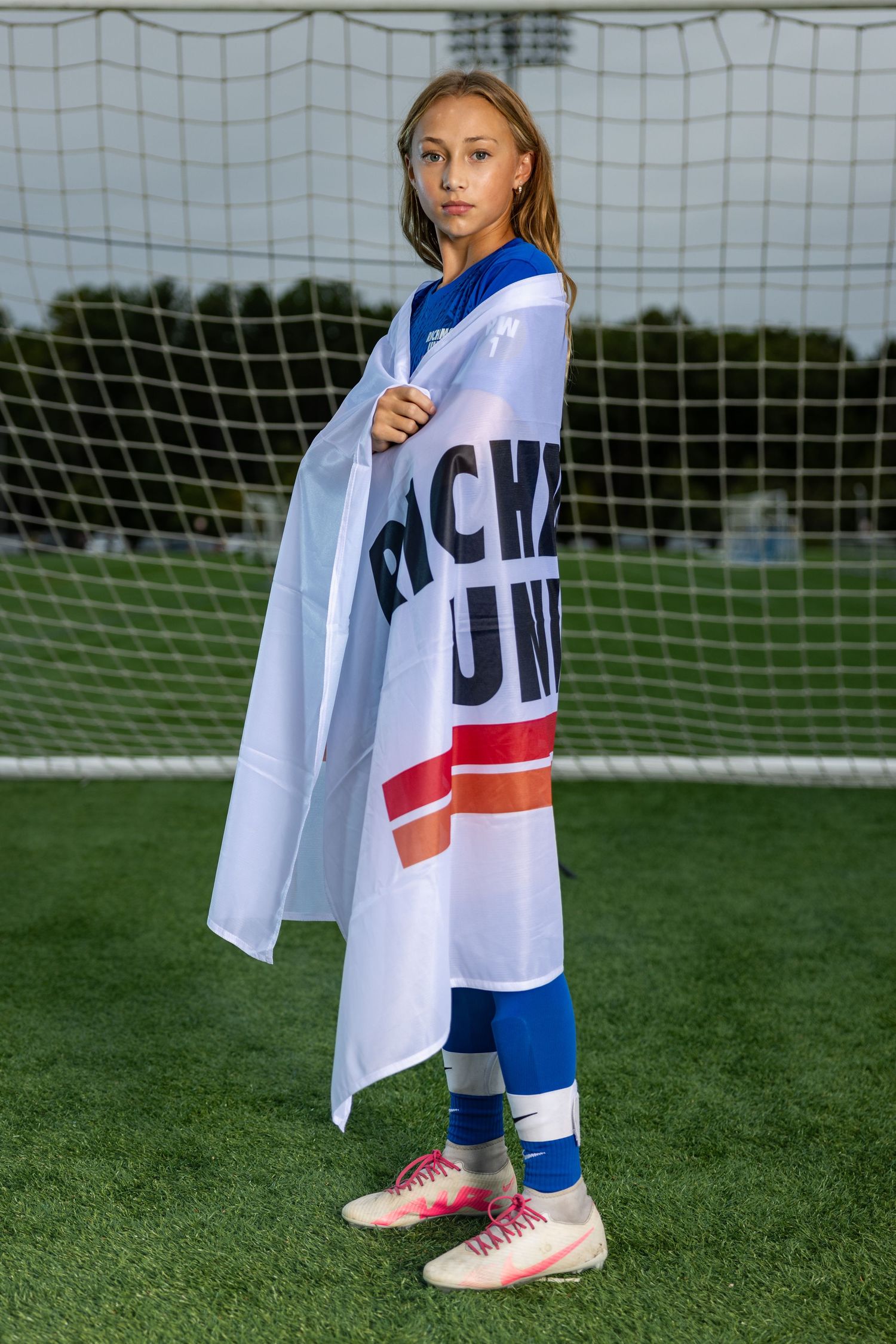 Soccer player wearing white jersey and blue pants poses with team flag on grass field near goal net.