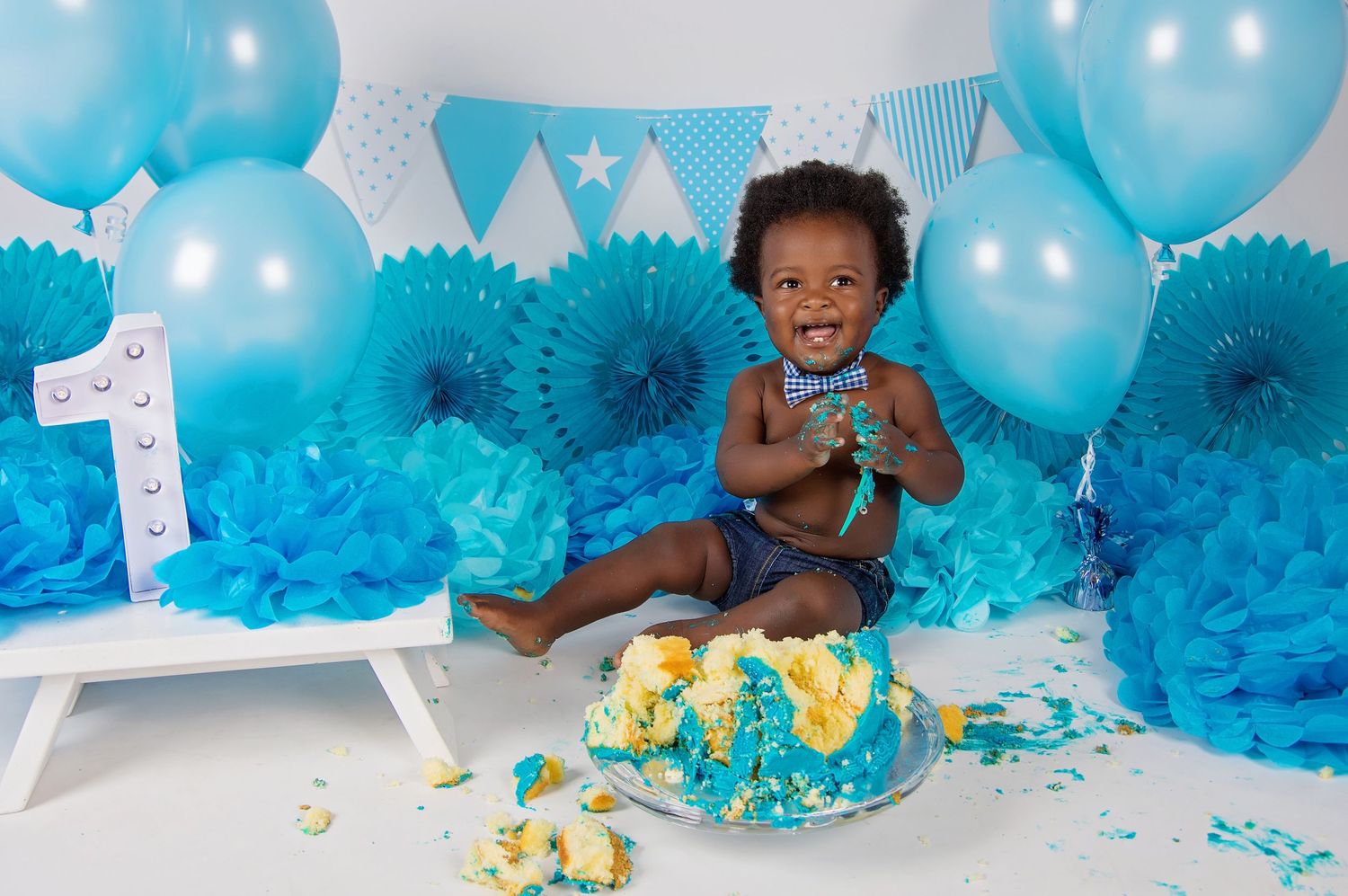 baby boy surrounded by blue decorations and balloons eating cake and laughing