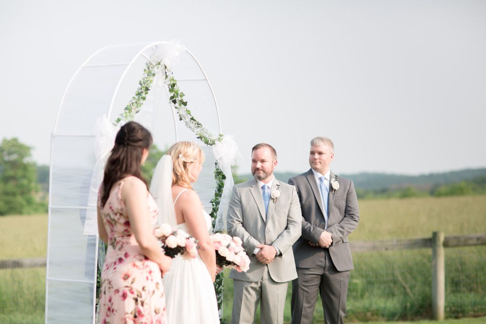 Outdoor wedding ceremony under white arch decorated with greenery in rustic countryside setting.