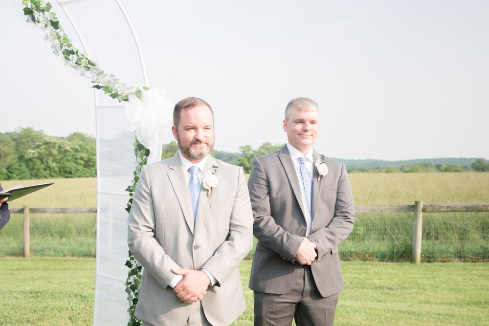 Groomsmen in light gray and charcoal suits stand by white wedding arch with greenery.