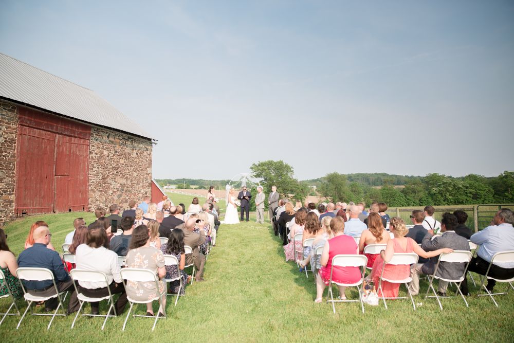 Rustic outdoor wedding ceremony setup with red barn and rolling hills in background.