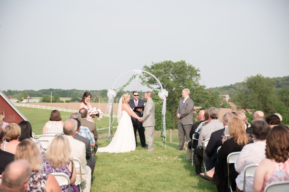 Couple exchanges vows under white floral arch at outdoor farm wedding ceremony.