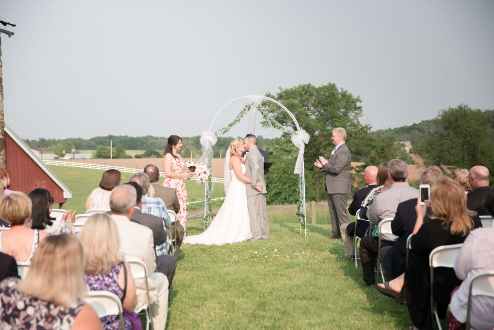 Wedding ceremony in progress under white arch with red barn and countryside views.