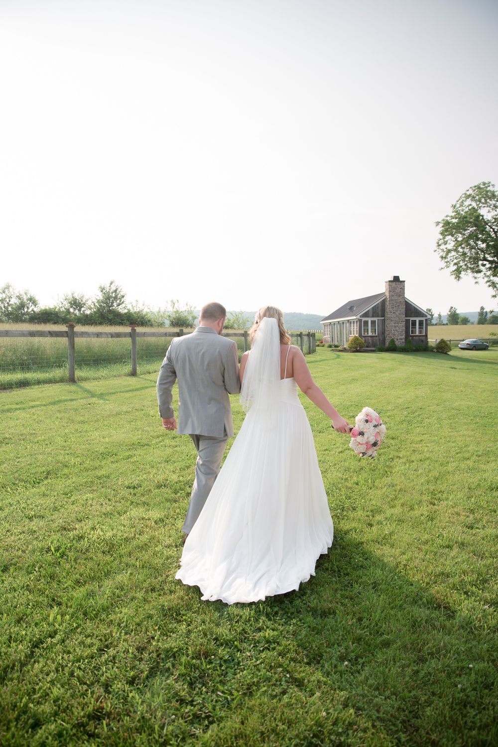 Newlyweds walk across grassy field towards rustic farmhouse after ceremony.