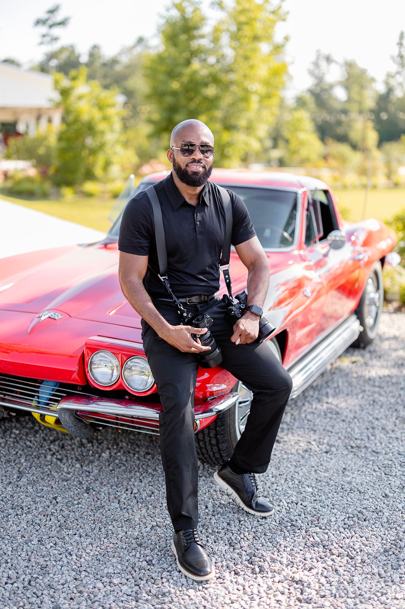 Professional photographer sits on vintage red Corvette wearing all black attire.