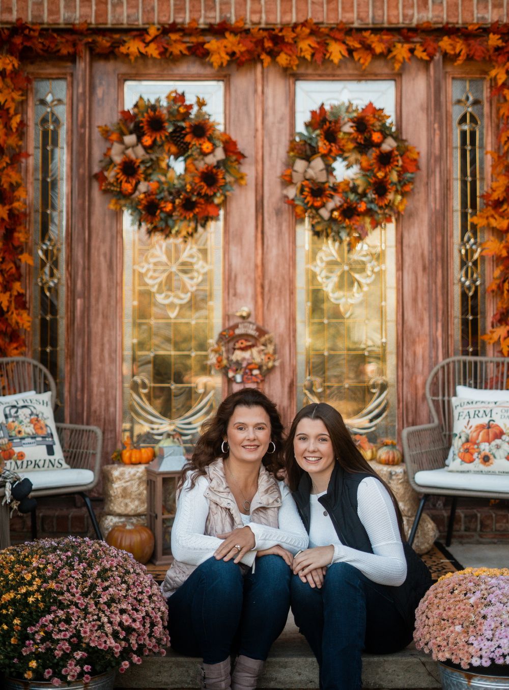 Two people sitting on porch steps with autumn wreaths and decorations in background.