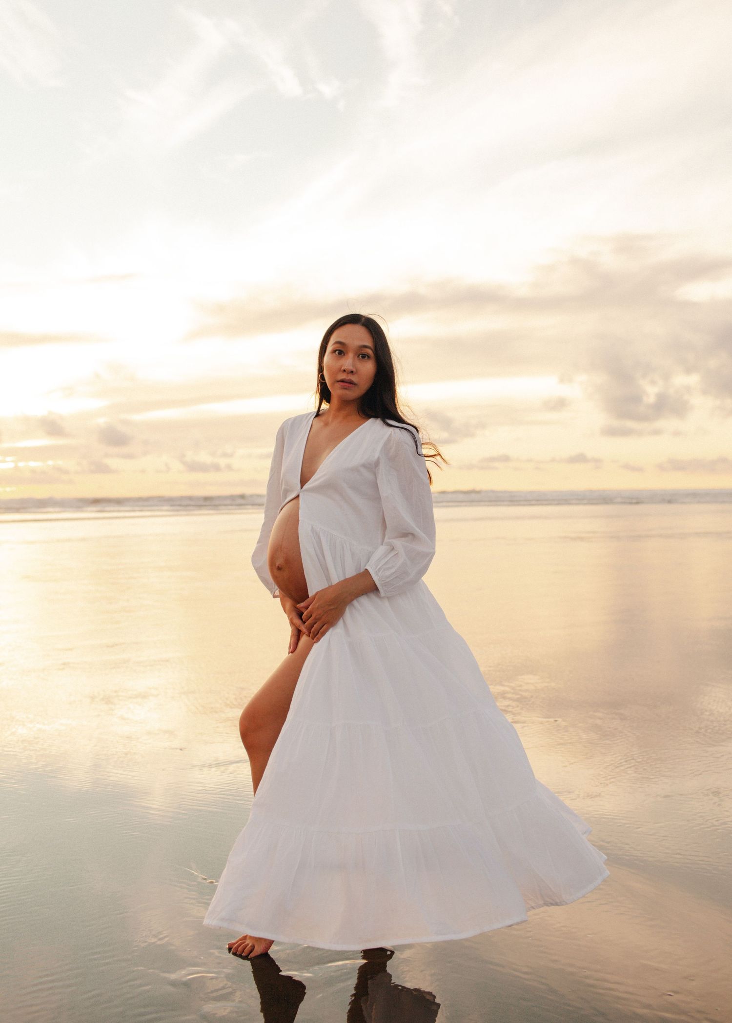 A person in a flowing white dress twirls and poses on a beach at sunset.