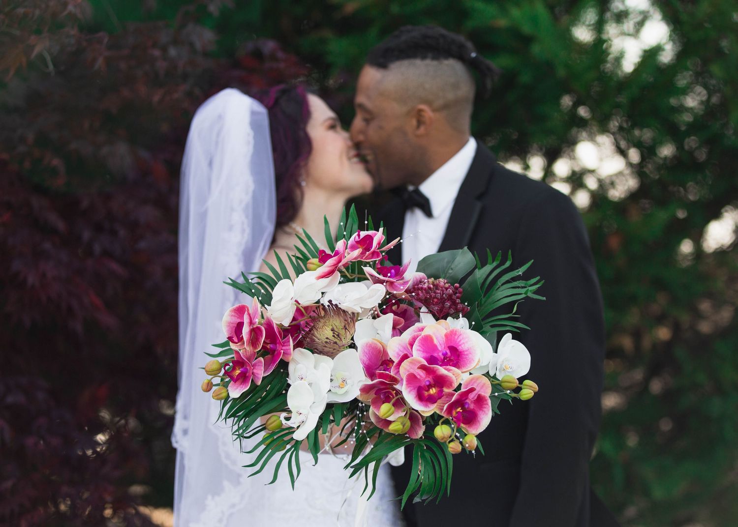 Wedding couple sharing a kiss with bride holding a vibrant tropical floral bouquet in lush garden.