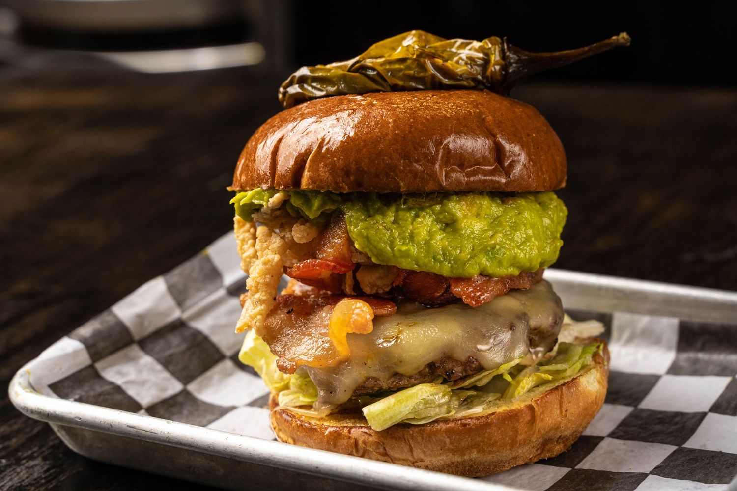 Gourmet burger with melted cheese, guacamole and crispy onions on checkered paper.
