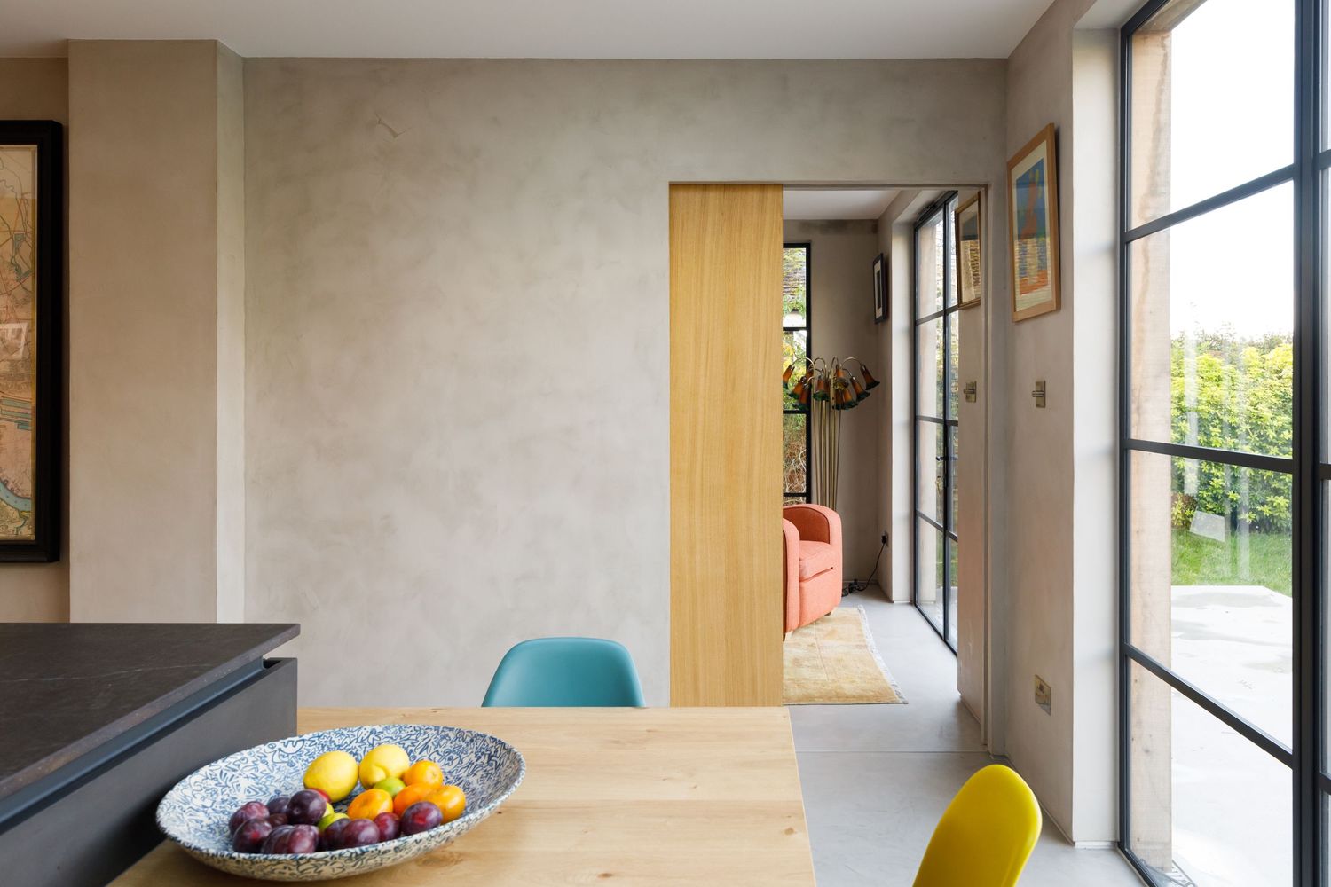 Bright dining area with colorful chairs and fruit bowl on wooden table next to large windows taken during an interiors photoshoot.