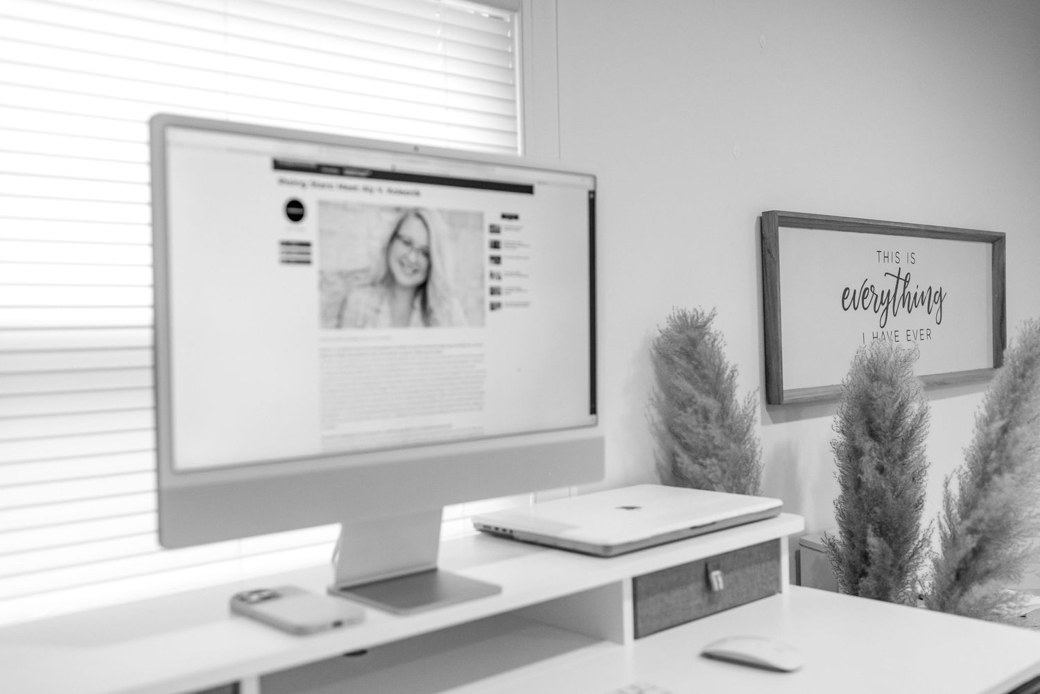 Black and white photo of iMac on white desk with pampas grass decor and framed motivational sign.