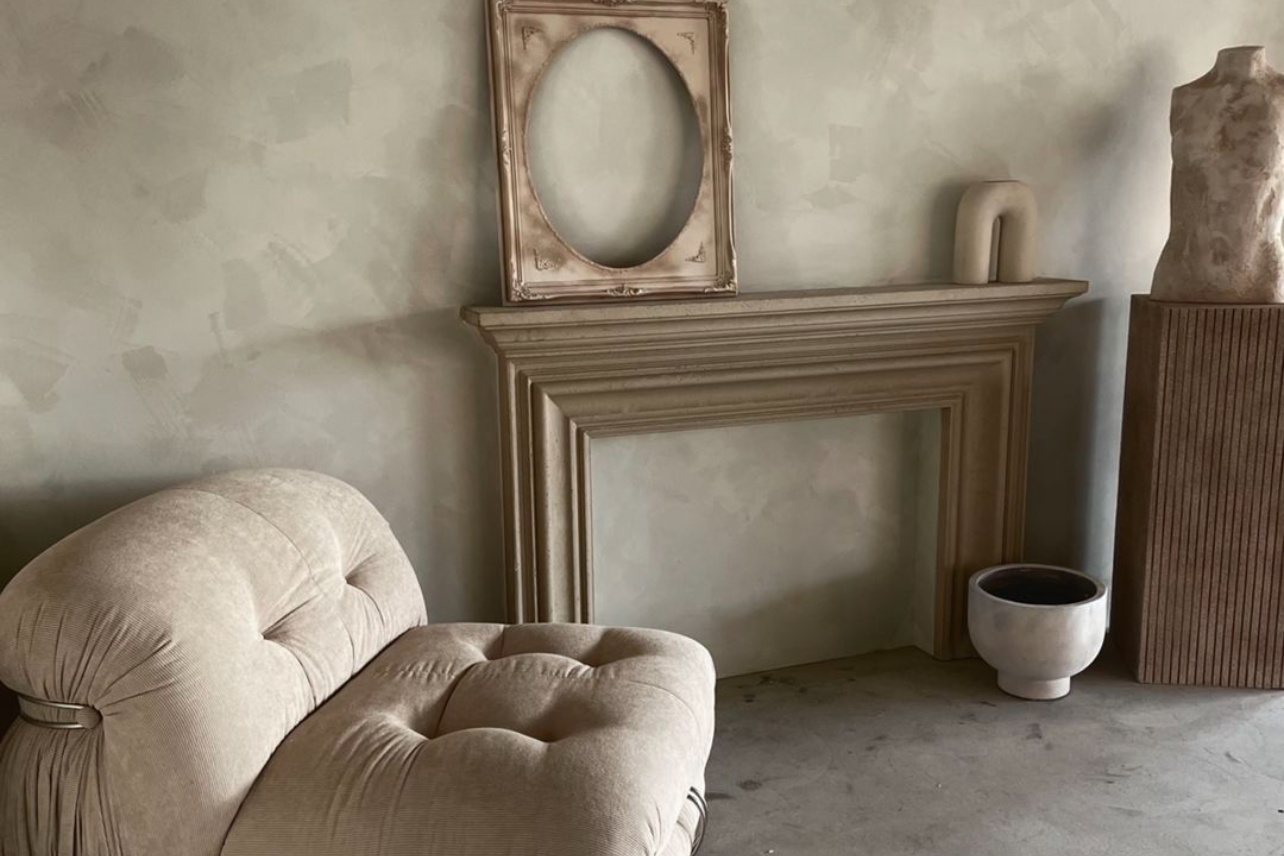 Minimalist living space with beige tufted chair and empty vintage frame above fireplace mantel.