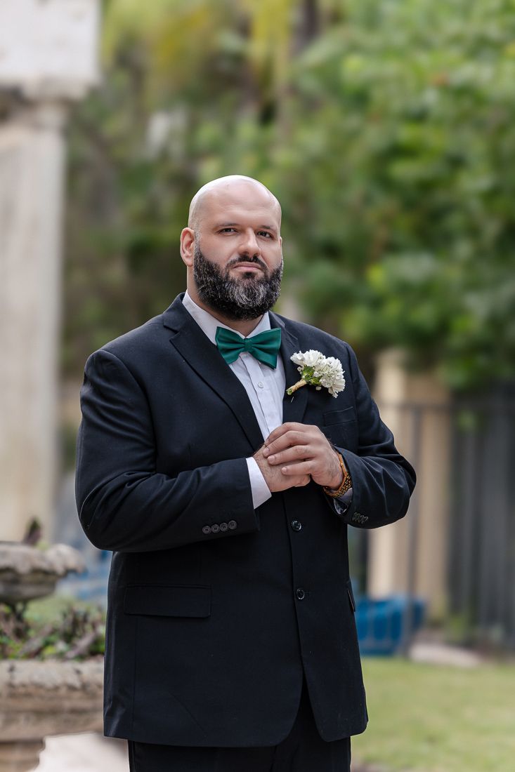 Groom in black tuxedo with teal bow tie and boutonniere adjusts his cuffs during wedding day.
