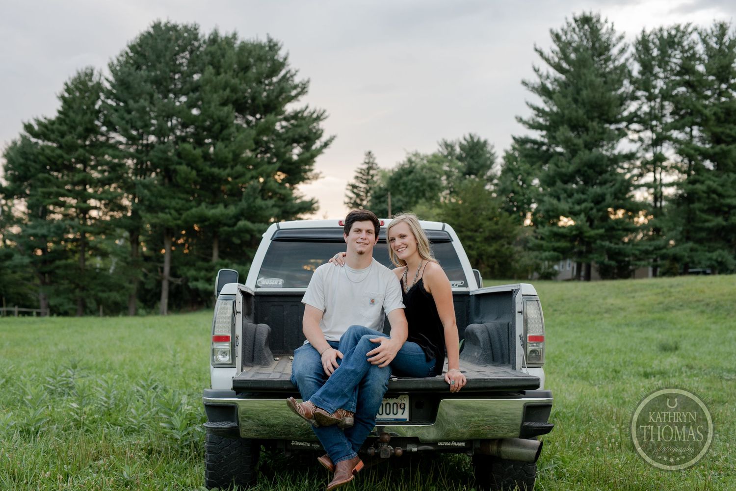A couple sits together in the back of a pickup truck with evergreen trees behind them.