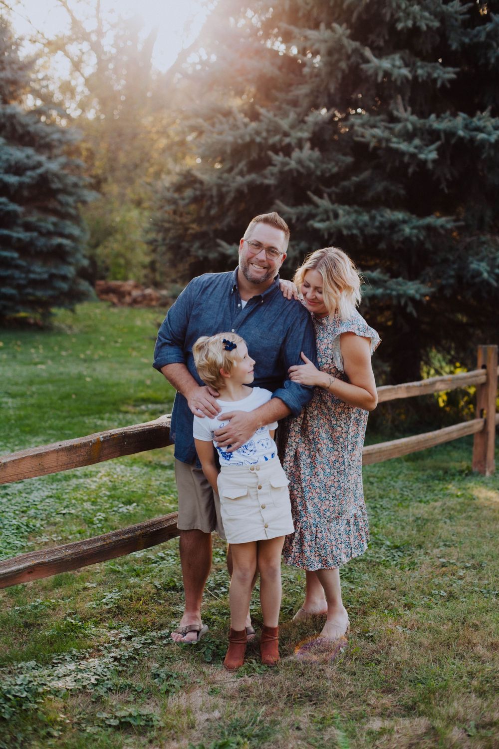 A sweet family moment captured at sunset near a rustic wooden fence with evergreen trees in the background.
