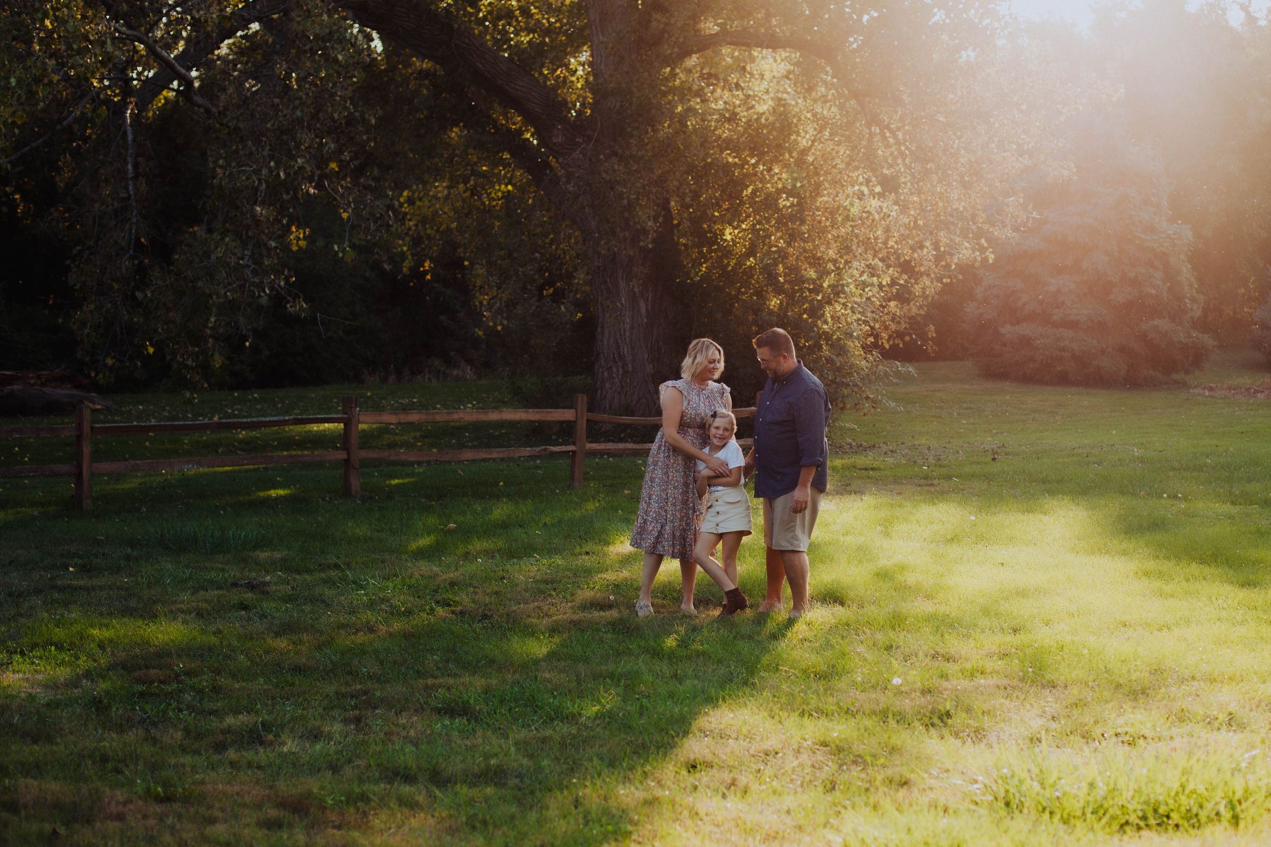 Three people share a tender moment while sitting on a wooden fence at sunset in a park setting.