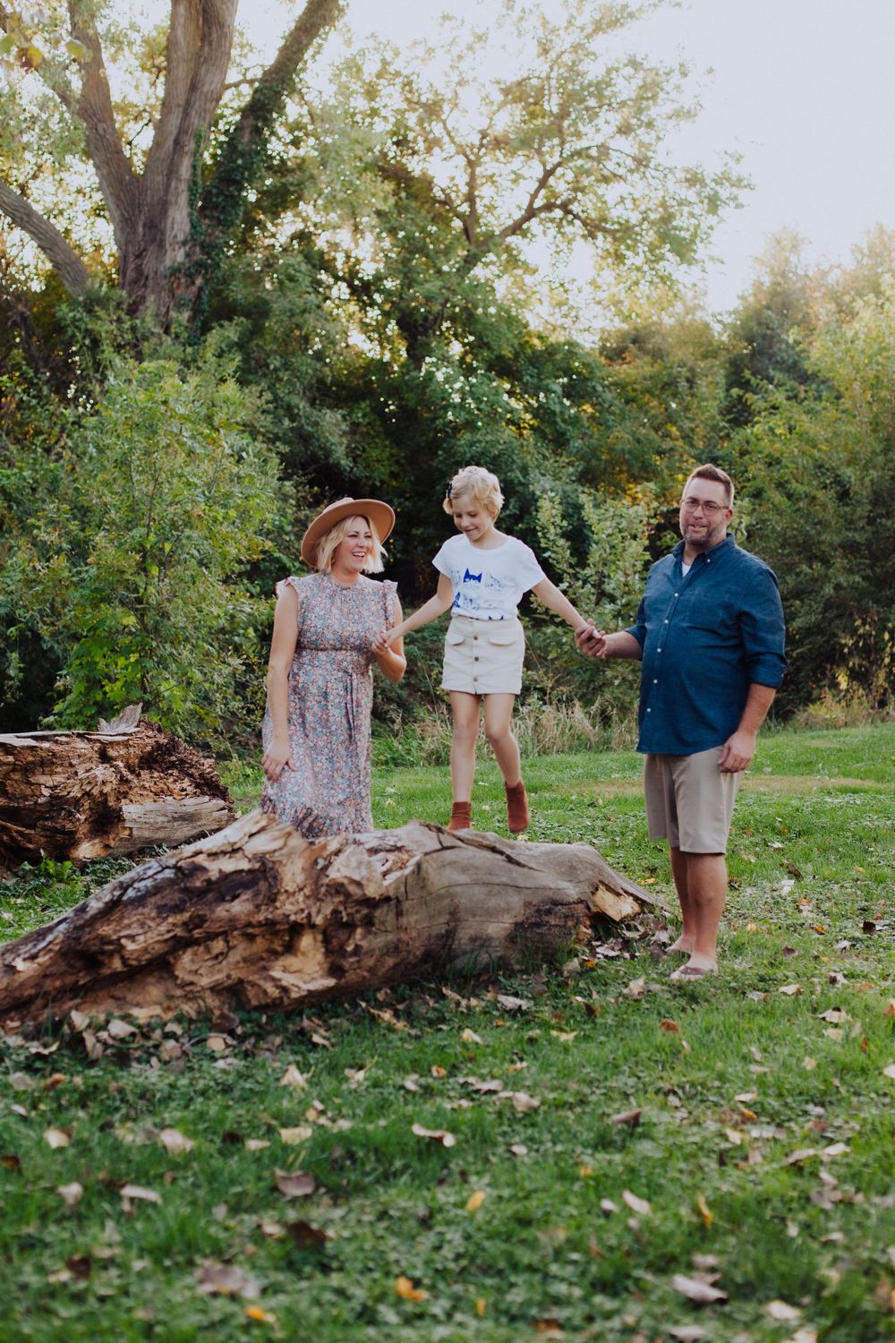 A playful family scene unfolds around a large fallen tree trunk in a sunlit garden setting.
